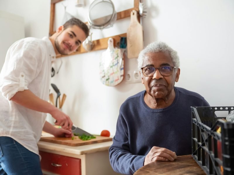 Young caregiver preparing food while an older man looks on at home